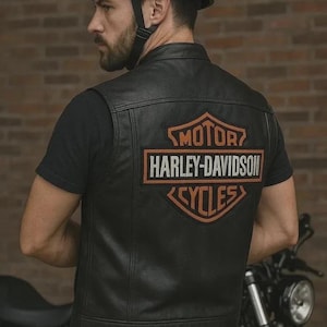 Könnte beinhalten: Schwarze Motorradweste aus Leder mit dem Harley-Davidson-Logo auf dem Rücken. Die Weste wird über einem schwarzen T-Shirt getragen. Ein schwarzer Helm wird getragen. Ein Motorrad ist im Hintergrund teilweise sichtbar.