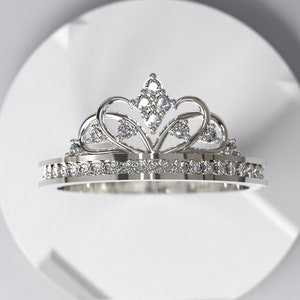 Può includere: Un anello d'argento con un design a corona e pietre trasparenti. La corona è realizzata con un'elaborata lavorazione in metallo e ha una punta appuntita.