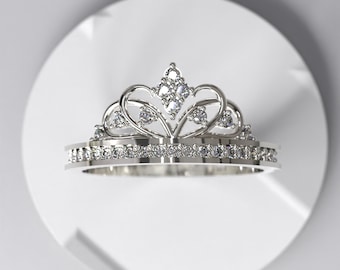 May include: A silver tiara ring with a crown design. The ring features a band encrusted with small, clear gemstones and a crown-shaped top adorned with larger, sparkling stones. The crown has heart-shaped accents and a central diamond cluster.