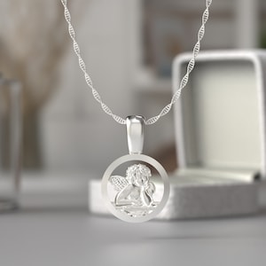 May include: A silver pendant necklace featuring a detailed angel design within a circular frame. The angel is depicted with wings and is resting its head on its hand. The necklace has a delicate, twisted chain.