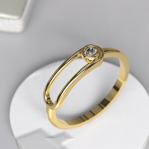May include: A gold ring with a single, clear diamond set in a bezel setting. The ring has a unique, open design with a curved band.