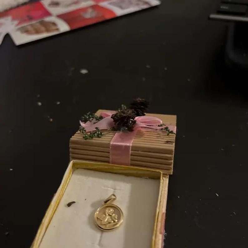 Peut inclure: Un petit pendentif d'ange en or dans une bo&icirc;te cadeau avec un ruban rose et des accents de pommes de pin.