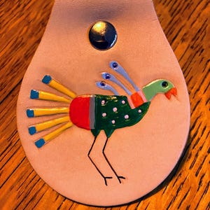 Könnte beinhalten: Ein hellbrauner Leder-Schlüsselanhänger mit einem bemalten Vogeldesign. Der Vogel hat einen grünen Kopf, einen roten und grünen Körper sowie gelbe und blaue Schwanzfedern. Ein schwarzer Druckknopf sichert den Schlüsselanhänger.
