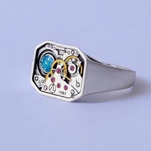May include: A silver ring with a square face featuring a detailed clockwork design with a blue and pink gemstone. The ring is set against a white background.