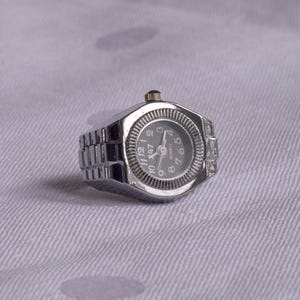 May include: A silver ring watch with a silver band and a black face. The watch face has white numbers and hands. The watch is on a white fabric background.