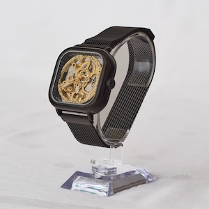 Puede incluir: Un reloj esqueleto negro con una esfera cuadrada y una correa de malla. El reloj presenta un intrincado movimiento dorado visible a través de la esfera transparente. El reloj se exhibe sobre un soporte de acrílico transparente.