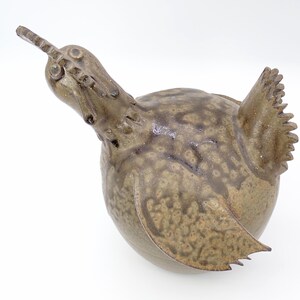 Può includere: Una scultura in ceramica di un uccello maculato di marrone e bianco con la testa inclinata all'indietro. L'uccello ha un becco lungo e appuntito e una piccola coda.