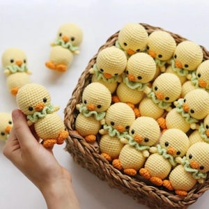 Può includere: Un cesto pieno di pulcini gialli all'uncinetto con accenti verdi e arancioni. I pulcini sono disposti in cerchio, con alcuni che spuntano dal cesto.