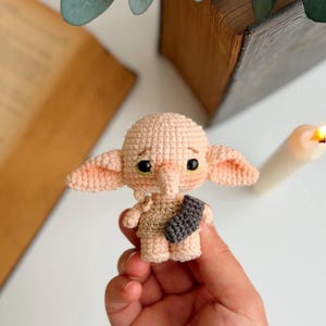May include: A small, beige crochet doll featuring large pointed ears, a long nose, and black bead eyes. The doll wears a tan tunic and holds a small, dark gray crocheted sock across its chest, crafted with detailed yarn stitching.