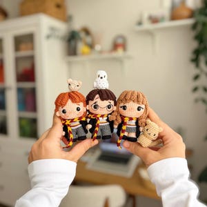 May include: Three small crochet dolls representing characters with brown, ginger, and light brown hair. They wear black robes, grey trousers or skirts, and scarves with red and yellow stripes. One doll has a white owl and another a small tan animal.