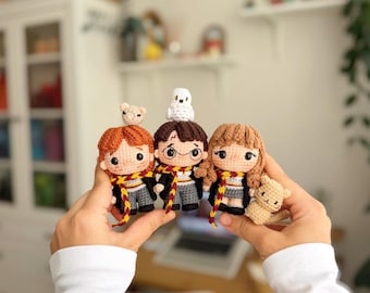 May include: Three small crochet dolls representing characters with brown, ginger, and light brown hair. They wear black robes, grey trousers or skirts, and scarves with red and yellow stripes. One doll has a white owl and another a small tan animal.