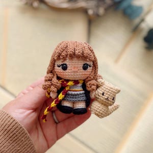 May include: A small crochet doll with curly brown hair, wearing a grey skirt, black shoes, and a scarf with red and yellow stripes. The doll is holding a small, light tan crocheted cat figure. The doll has large black eyes and stitched facial features.