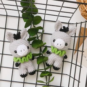 May include: Two grey crocheted donkey plushies with black eyes and green ruffled collars. The plushies are sitting on a black wire mesh surface.