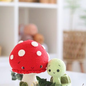 May include: A red and white crocheted mushroom with a smiling face and a green crocheted turtle with a smiling face. The mushroom has a white stem and green leaves at the base. The turtle is sitting next to the mushroom.