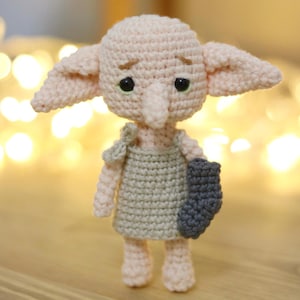 May include: A handmade crochet doll in the likeness of a character with large ears, a long nose, and large eyes. The doll is primarily light pink with a green and gray outfit and a gray sock. The doll is standing upright.