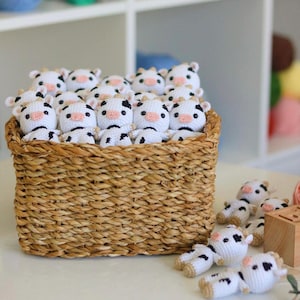 May include: A wicker basket filled with crocheted cow plushies. The plushies are white with black spots and have pink noses.