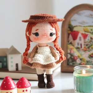 May include: A handmade crochet doll with red braids and a brown hat, wearing a white dress and brown boots. The doll is standing on a white surface, with miniature houses and a candle in the background. The doll is approximately 6 inches tall.