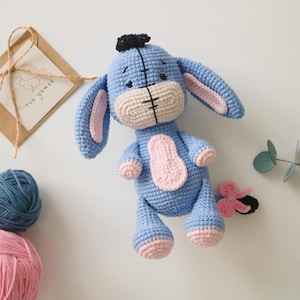 May include: A blue crocheted stuffed animal resembling a donkey with pink ears, hooves, and a tail. The animal has black eyes and a black nose.