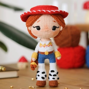 Puede incluir: Muñeca de ganchillo de un personaje que lleva un sombrero rojo, una camisa blanca con ribetes amarillos, vaqueros azules y polainas con estampado de vaca. La muñeca tiene el pelo naranja y una expresión amigable. Un juguete hecho a mano.