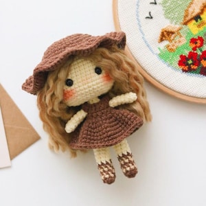 Puede incluir: Una muñeca de crochet con cabello castaño, un vestido marrón y un sombrero marrón. La muñeca lleva calcetines a rayas marrones y blancas.