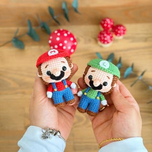 Puede incluir: Dos pequeñas muñecas de ganchillo que representan a Mario y Luigi. La muñeca de Mario lleva una gorra roja con una M, una camisa roja y un peto azul. La muñeca de Luigi lleva una gorra verde con una L, una camisa verde y un peto azul.