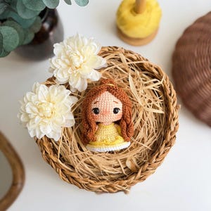 Op de afbeelding: Een kleine gehaakte pop met bruin garen haar in een paardenstaart, gekleed in een gele jurk met een witte rand. De pop heeft zwarte kraaloogjes en roze wangen en zit in een ronde gevlochten mand gevuld met stro-achtig materiaal.