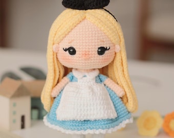 May include: A handmade crochet doll of a character with blonde hair, a black bow, and a blue dress with a white apron. The doll has black eyes and is standing on a white surface. The doll is approximately 6 inches tall.