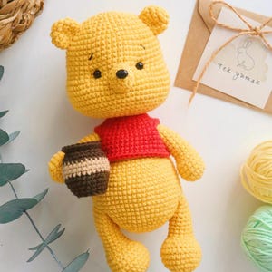 Puede incluir: Un peluche de Winnie the Pooh tejido a crochet sosteniendo un tarro de miel. El juguete es amarillo con una camisa roja y ojos marrones. El fondo incluye un sobre marrón con el texto "Tek yumak" y una planta verde.