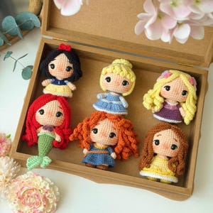 Puede incluir: Seis pequeñas muñecas de ganchillo dispuestas en una caja de madera. Las muñecas tienen pelo de lana en negro, rubio, rojo y marrón, y visten trajes coloridos que incluyen vestidos y una cola de sirena. Cada muñeca tiene grandes ojos negros.