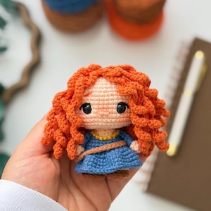 Peut inclure: Une petite poupée au crochet avec des cheveux bouclés orange vif, un visage couleur pêche clair aux yeux noirs, et une robe bleue avec une encolure jaune et une ceinture marron. La figurine est réalisée avec des mailles détaillées et compactes.
