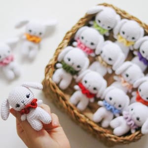 May include: A basket filled with crocheted white bunny plushies with colorful accents. The bunnies have different colored bows and collars.