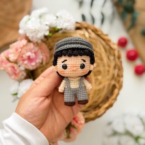 May include: A small crochet doll featuring a person with dark hair wearing a grey flat cap, a cream-colored shirt, and grey overalls with brown shoes. The doll has black eyes, rosy cheeks, and a neutral expression, crafted with detailed yarn stitching.