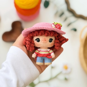 Pode incluir: Uma pequena boneca de crochê com cabelo encaracolado castanho-avermelhado, usando um chapéu de sol rosa adornado com um morango minúsculo, uma camisa branca de manga comprida com listras horizontais vermelhas e verdes e calções azul-claros.