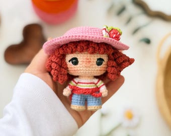Puede incluir: Una pequeña muñeca de ganchillo con pelo rizado de color marrón rojizo, que lleva un sombrero de sol rosa adornado con una fresa diminuta, una camiseta blanca de manga larga con rayas horizontales rojas y verdes, y pantalones cortos azul claro.