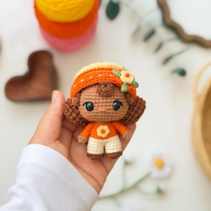 May include: A small crochet doll with brown hair, wearing an orange hat with a small flower, an orange long-sleeved shirt with a white flower detail, and white pants. The doll has large black eyes and is crafted with visible yarn stitches.