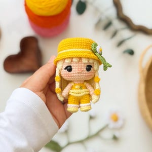 May include: A small crochet doll featuring light blonde hair with yellow accents, wearing a bright yellow bucket hat adorned with a green leaf and white flower detail, and a matching yellow outfit consisting of a top and shorts with white trim.
