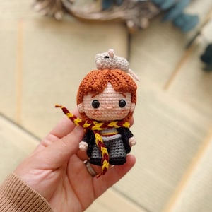 May include: A small crochet doll featuring a character with ginger hair, wearing a black robe over a grey outfit, and a yellow and maroon striped scarf. A tiny light grey crocheted rat sits on top of the character's head. The doll has black eyes.