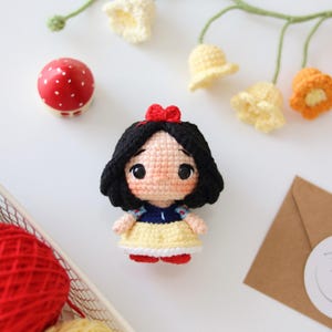 Puede incluir: Una pequeña muñeca de ganchillo con cabello negro y un lazo rojo, un corpiño azul y una falda amarilla. La muñeca tiene grandes ojos negros y piel clara. Está acompañada por un hongo rojo, flores de ganchillo y ovillos de lana.