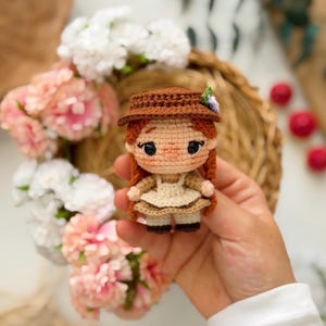 May include: A small crocheted doll with red yarn hair styled in two braids, wearing a brown hat with a small purple flower, a beige dress with a white apron, and dark brown shoes. The doll has large black eyes and a light peach-coloured face.