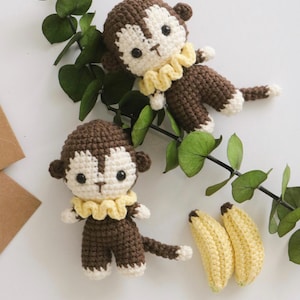 May include: Two brown crocheted monkey toys with yellow collars and two yellow crocheted bananas. The monkeys are sitting on a branch with green leaves.