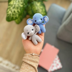 May include: Two crocheted elephant toys, one gray and one light blue, are held in a person's hand. The toys have embroidered eyes and a small trunk.