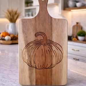 May include: A wooden cutting board with a handle, featuring a carved pumpkin design. The board is made of light and dark brown wood, perfect for fall-themed kitchen decor. The pumpkin carving adds a seasonal touch to the kitchen.