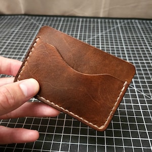 May include: A brown leather card holder with a white stitch detail. The card holder has a single pocket for cards.