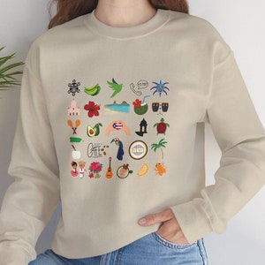 Woman wearing a sand-colored sweatshirt featuring Puerto Rican symbols, including the Boricua flag, Taino designs, and Boricua emblems, showcasing cultural pride.
