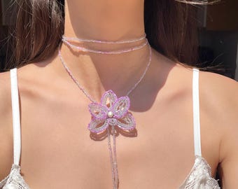 May include: A beaded necklace featuring a large, lavender flower pendant with a pearl center. The necklace has a delicate, layered design with strands of small beads in shades of pink and white. The jewelry is a floral accessory.