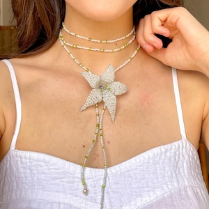 May include: A white beaded necklace with a large white flower pendant. The necklace is layered and has green beads interspersed throughout.