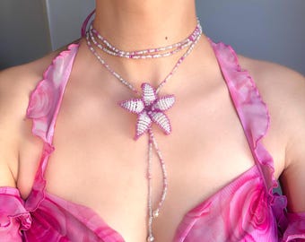 May include: A beaded necklace with a large, flower-shaped pendant. The necklace features white and pink beads, with a long beaded strand ending in two small, pearly beads. The model is wearing a pink dress with a floral pattern.