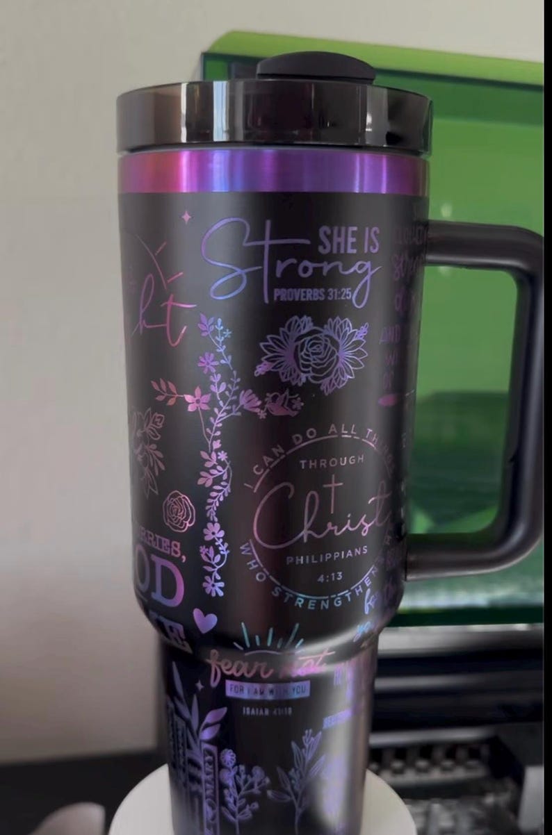 May include: Black stainless steel tumbler with a floral design and inspirational quotes. The quotes include "She is Strong, Proverbs 31:25", "I can do all things through Christ, Philippians 4:13", and "Fear not, for I am with you, Isaiah 41:10".