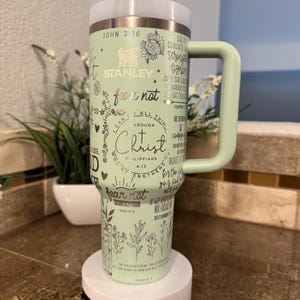 May include: A green stainless steel tumbler with a white lid and a black straw. The tumbler has a variety of Christian-themed quotes and designs, including the words "fear not" and "I can do all things through Christ". The tumbler also features the Stanley logo.