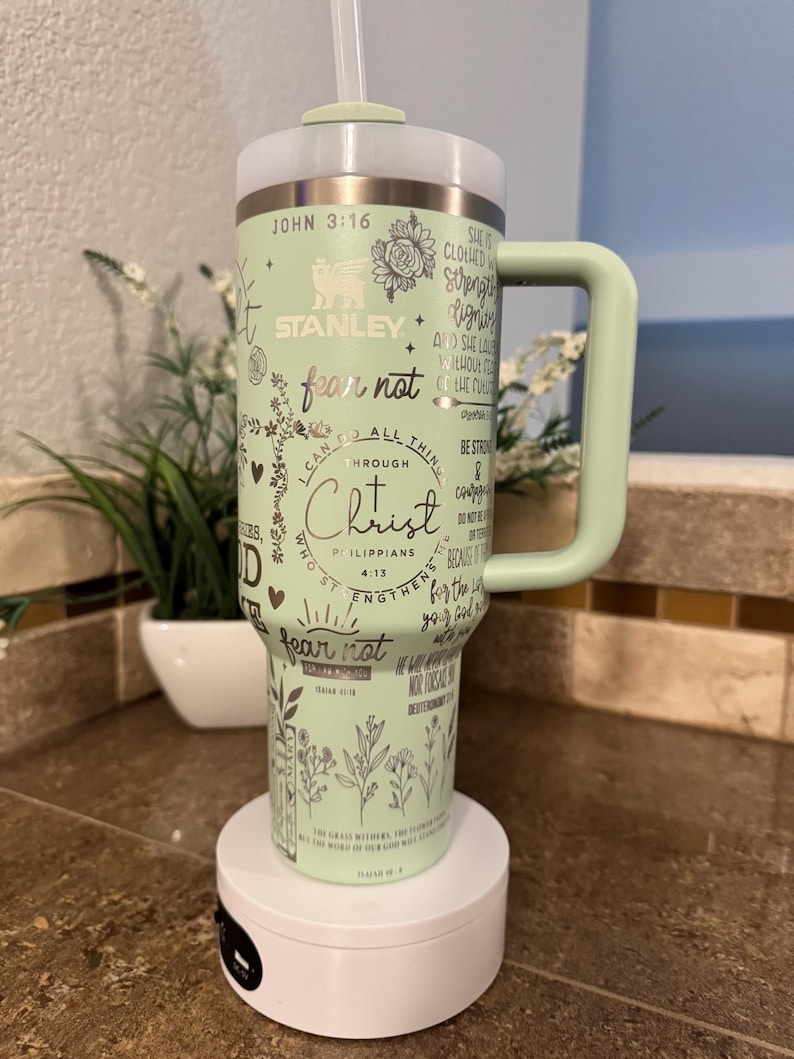 May include: A green stainless steel tumbler with a white lid and a black straw. The tumbler has a variety of Christian-themed quotes and designs, including the words "fear not" and "I can do all things through Christ". The tumbler also features the Stanley logo.